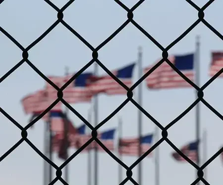Blurred American flags behind a chain-link fence outdoors.